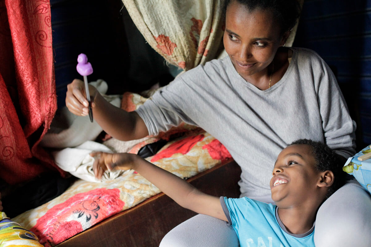 Abdi sitzt im Schoß seiner Mutter. Sie trägt ein graues T-Shirt und eine graue Leggings, er ein blaues Shirt. Der Bub zeigt mit einem Lächeln uaf einen Stift mit einer lila Plastikente, den seine Mutter hochhält.