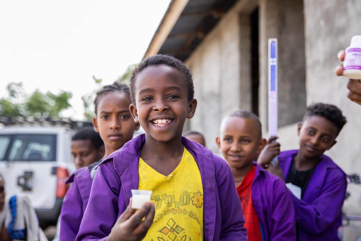 Auf dem Bild sind Schulkinder zu sehen. Sie haben eine dunkle Hautfarbeund tragen lila Schuljacken. Ein Junge steht ganz vorne und grinst in die Kamera. In der Hand hält er die Dosis eines flüssigen Antibiotikums gegen die Augeninfektion Trachom.