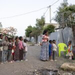 Auf dem Foto stehen mehrere Kinder auf einer Straße Schlange, um von einem sitzenden Gesundheitshelfer Medikamente zu erhalten.