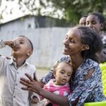 Auf dem Foto trinkt ein Junge Medikamente aus einem kleinen Becher, während seine Mutter lächelnd zusieht.