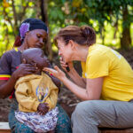 Auf dem Bild ist die Ärztin Dr. Irene Ruhswurm zu sehen. Sie sitzt auf einer Holzbank und untersucht gerade die Augen eines Kleinkindes. Das Kleinkind sitzt auf dem Schoß seiner Mutter. Das Bild wurde im afrikanischen Staat Uganda im Dorf Kikubo aufgenommen.