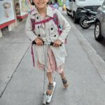 Auf dem Bild ist ein 9-jähriges Mädchen auf dem Weg in die Schule zu sehen. Sie steht auf einem Scooter und hat ihren Schulrucksack an. Sie lächelt und freut sich auf den Schulanfang.