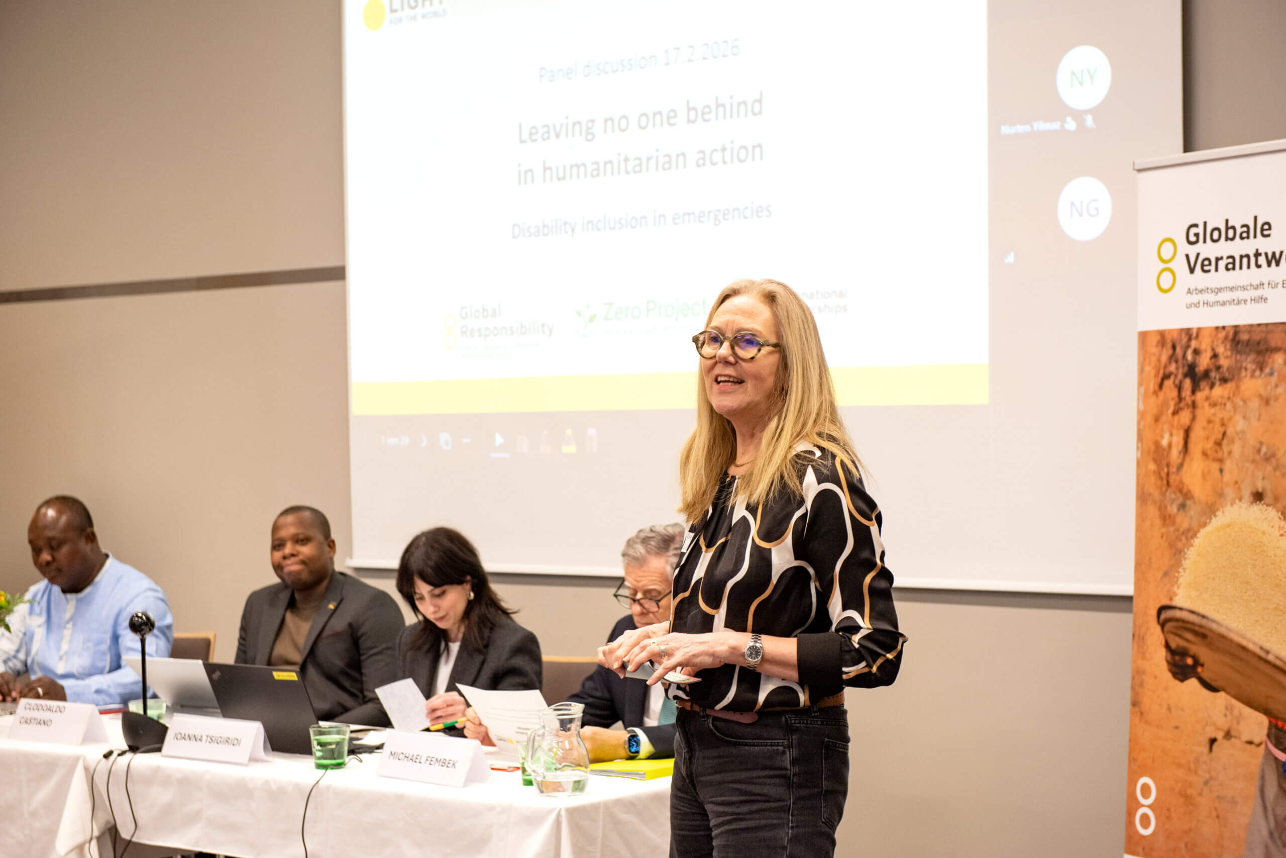 On the picture you can see a woman in her middle age. She wears a black pants and a black blouse with white spots on it. She wears glasses and has midlong hair. She is currently speaking to an audience at a panel discussion around inclusive humanitarian action. The woman is the CEO of Light for the World.