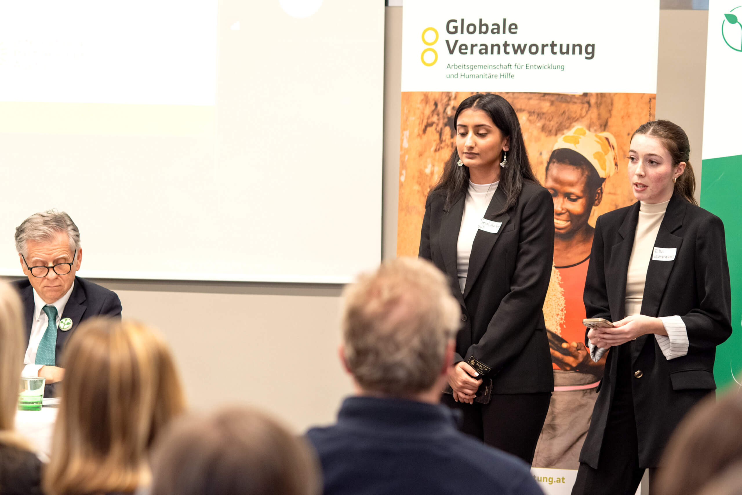 On the picture you can see two woman standing in front of an audience and talking to them. They are speakers during a panel discussion around inclusive humanitarian action. They are both wearing a black suite and a white blouse.