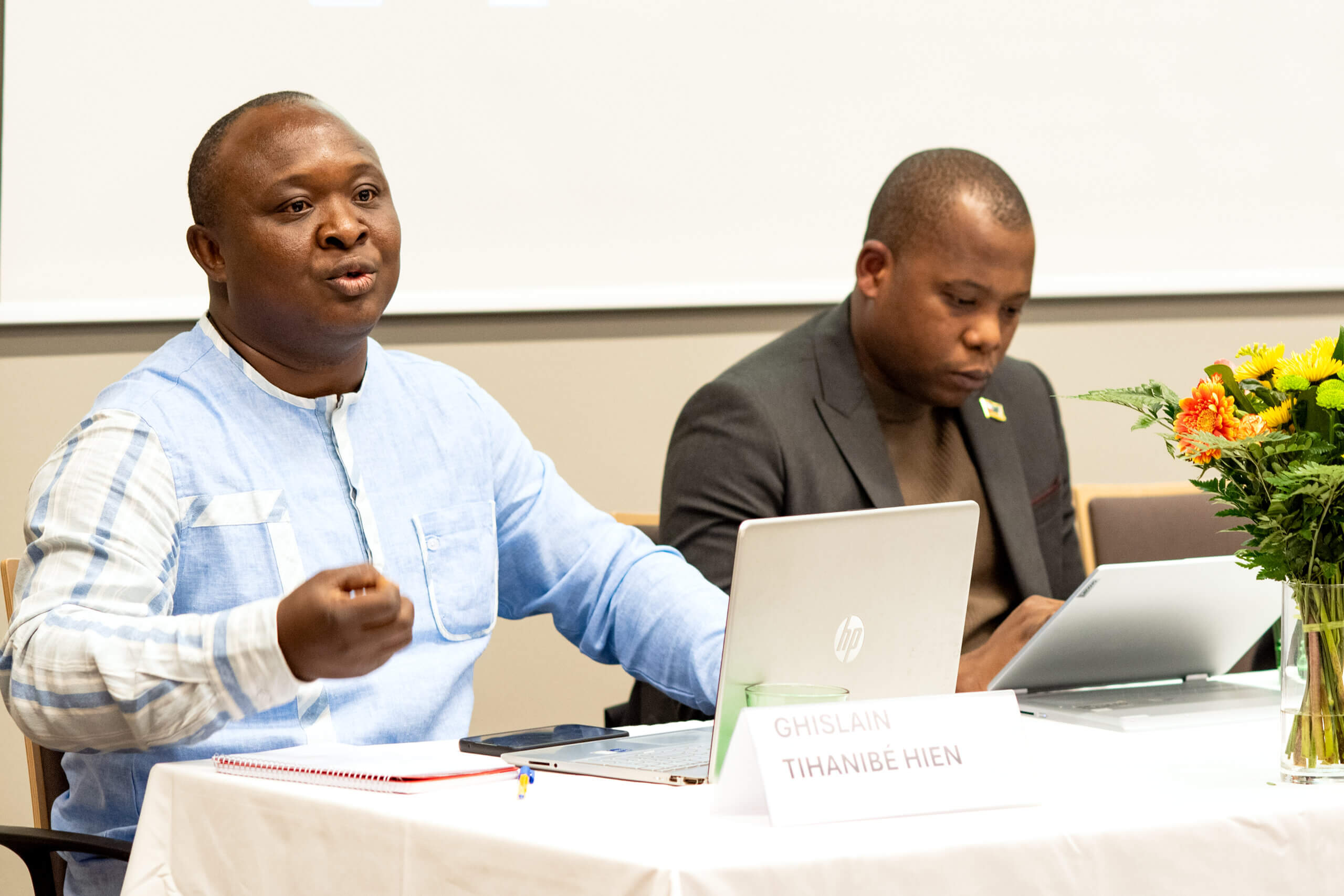 On the picture you can see a man who is sitting on a table. He is currently speaking to a audience about inclusive humanitarian action. Some documents are lying in front of him.