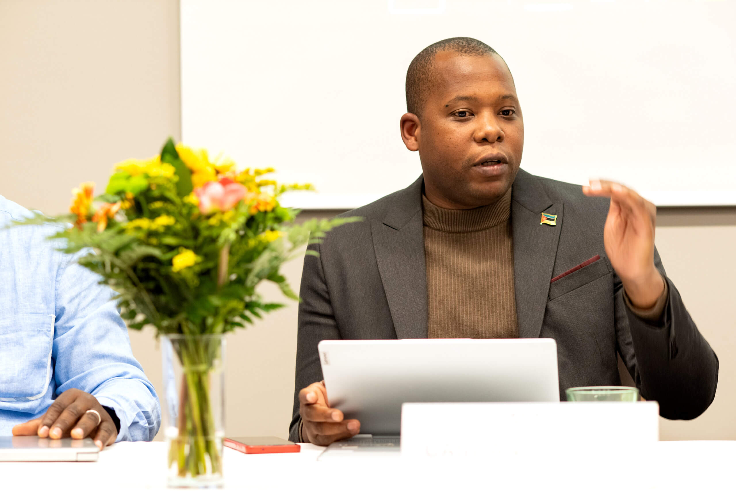On the picture you can see a man wearing a suite. He is sitting on a table and obviously talking. He is talking about inclusive humanitarian action. On the table you can see some documents and a bouqet of flowers.