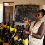 Der Bildausschnitt zeigt Esther, eine Inklusionsberaterin von Licht für die Welt, die vor einer Schulklasse gebärdet. Sie trägt ein beiges Gillet und hinter ihr sitzen zahlreiche Kinder in gelben T-Shirts am Boden und achten auf ihre Handbewegungen. Sie befinden sich in einem Flüchtlingscamp für Binnenvertriebene im Südsudan.