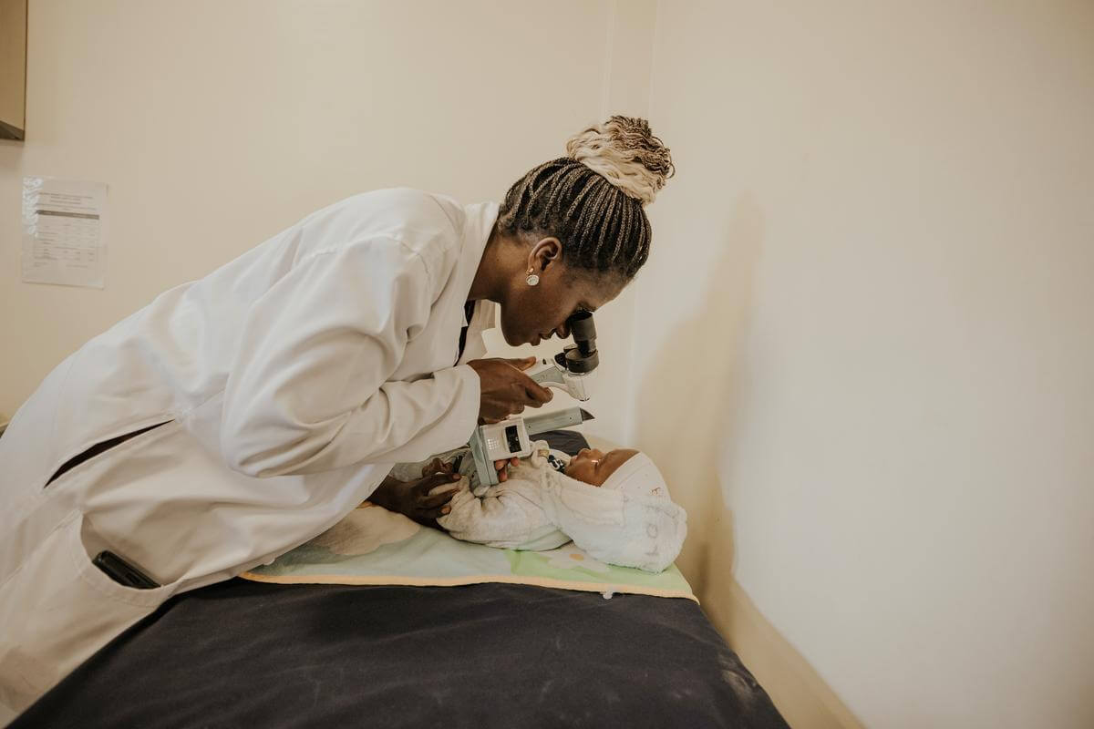 Eine schwarze Ärztin trägt einen weißen Kittel, während sie sich mit einem Untersuchungsgerät für das Auge über ihre Patienten beugt. Der Patient ist ein kleiner Junge, der wegen Grauem Star operiert wurde. Die Frau ist Ärztin mit Spezialgebiet Kinderaugenheilkunde.