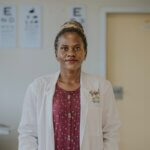 Eine schwarze Frau in einem weißen Kittel und einer roten Bluse blickt in die Kamera. Sie ist Kinderaugenärztin in einem Spital in Uganda.