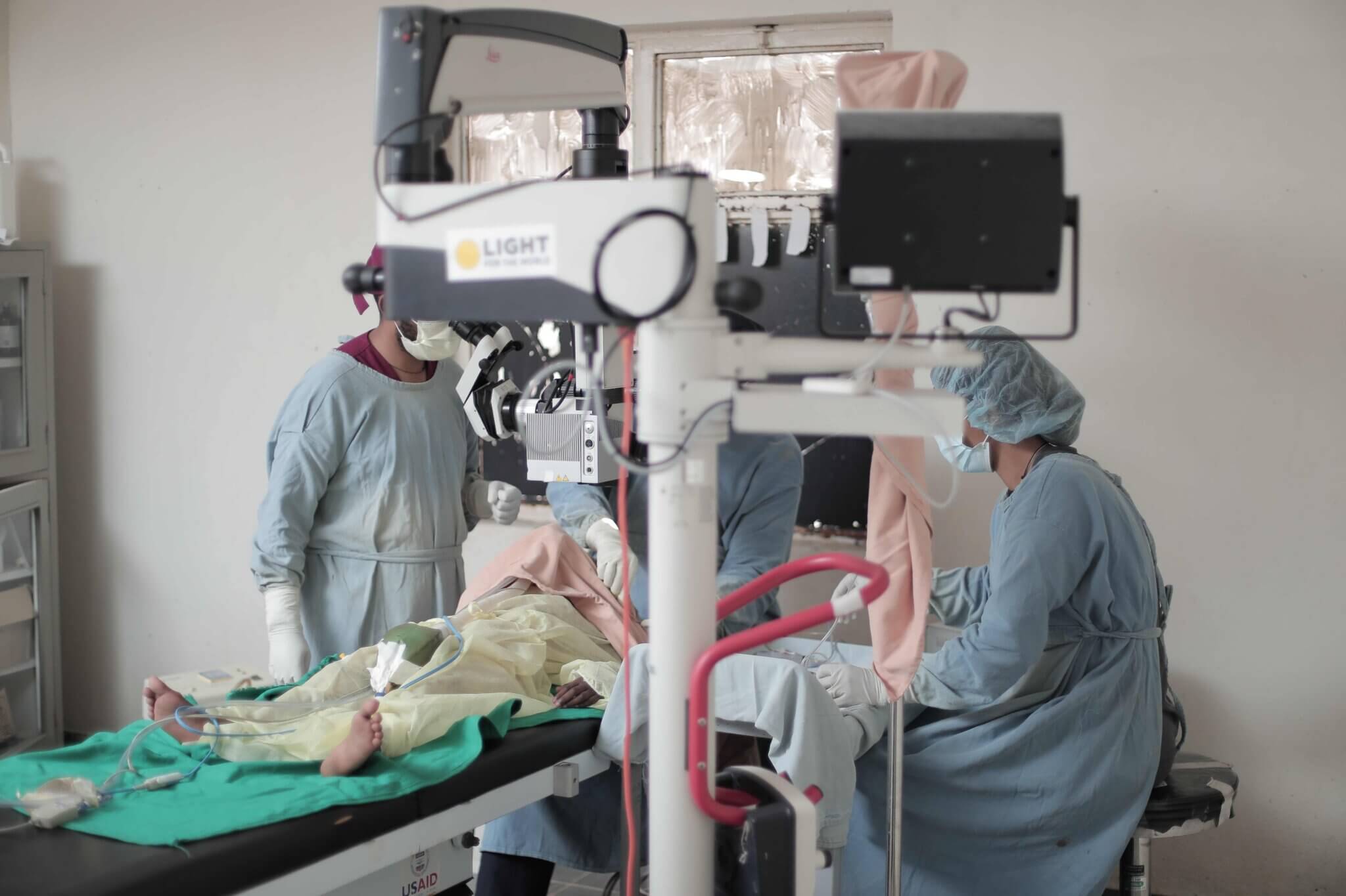 Auf dem Bild ist ein Operationssaal zu sehen. Auf dem Tisch liegt ein Kind, es wird am Auge operiert. Rundherum steht das Operations-Personal und führt den Eingriff durch.