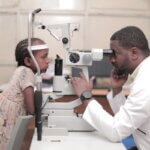 Ein junges Mädchen kniet auf einem Stuhl und hat ihren Kopf auf einem Untersuchungsgerät für Augen abgelegt. Ein Arzt untersucht sie gerade.