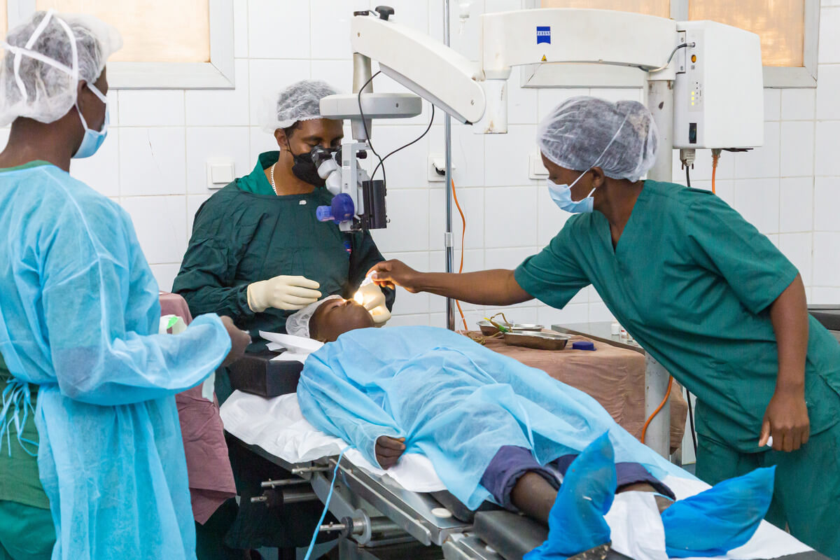 Auf dem Bild ist ein Operationssaal zu sehen. Ein Patient liegt auf dem Tisch, ein Augenarzt operiert in an seinem Auge. Um sie herum steht der Rest des Operationsteams.