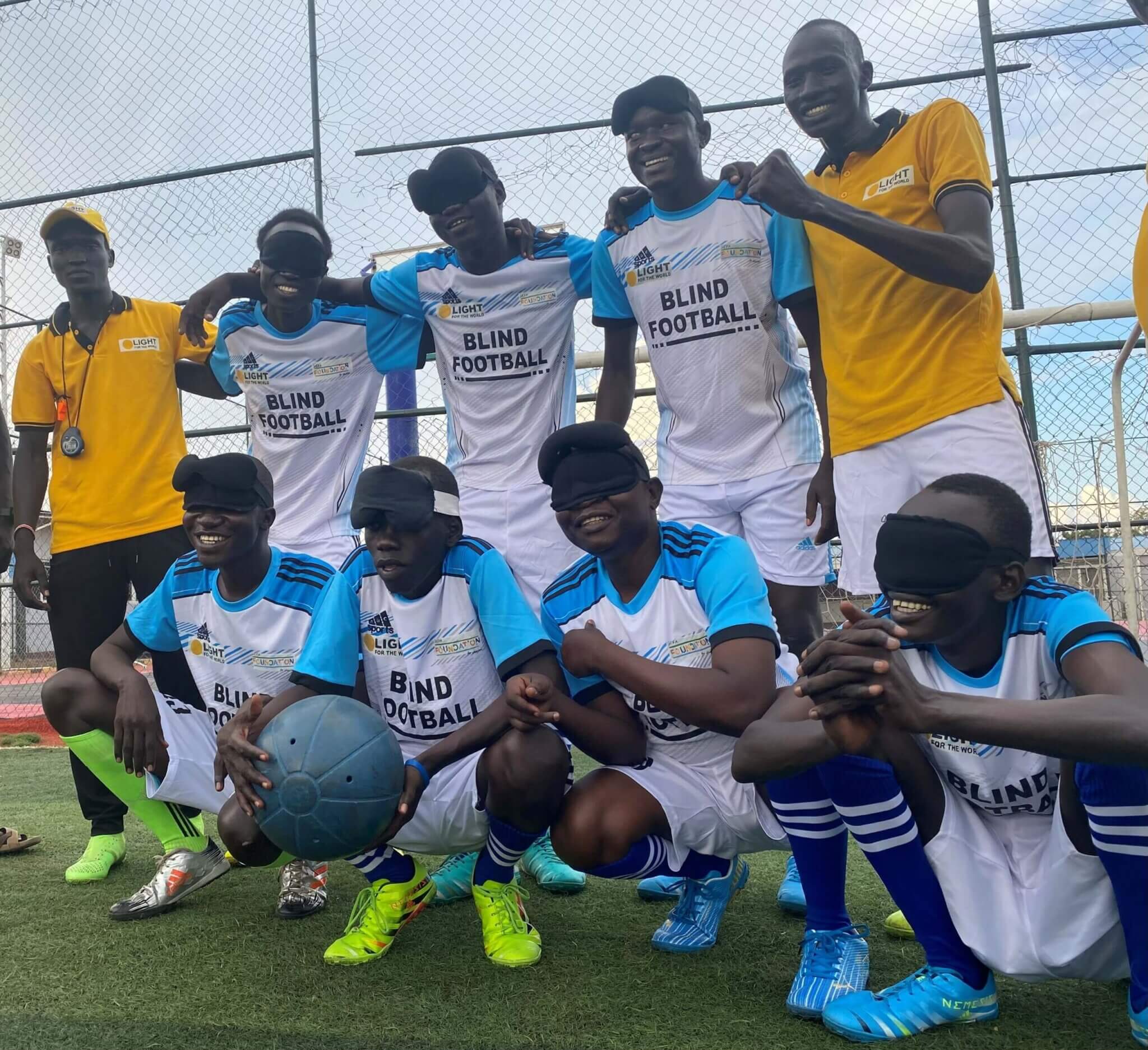 Zu sehen ist die Fußballmannschaft des blinden Fußballvereins im Südsudan. Die Spieler tragen Augenbinden und einer hält den Spielball. Simon, der Trainer, ist ebenfalls auf dem Bild.
