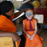 Ein Kind, das mit Albinismus lebt, sitzt in einer orangenen Schuluniform auf einem Holzsessel und blickt durch einen Versuchslinsenrahmen. Eine Frau in einem orangenen Shirt hält dabei ihren Kopf und findet gemeinsam mit ihr die passende Dioptrienstärke.