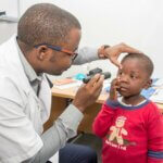 Der einzige Kinderaugenarzt aus Mosambik untersucht das Auge eines Jungen. Er leuchtet ihm mit einer Taschenlampe ins Auge, während das Kind die Haut seines Auges nach unten zieht und in das Licht der Taschenlampe blickt.