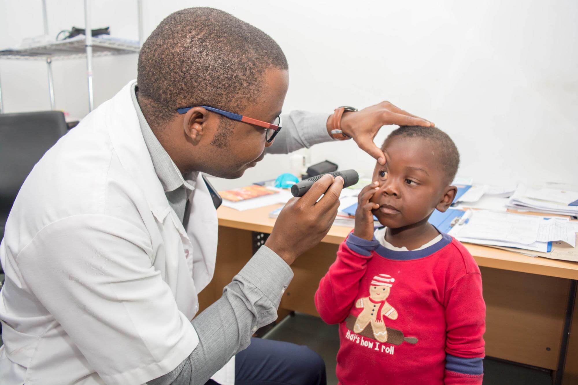 Der einzige Kinderaugenarzt aus Mosambik untersucht das Auge eines Jungen. Er leuchtet ihm mit einer Taschenlampe ins Auge, während das Kind die Haut seines Auges nach unten zieht und in das Licht der Taschenlampe blickt.