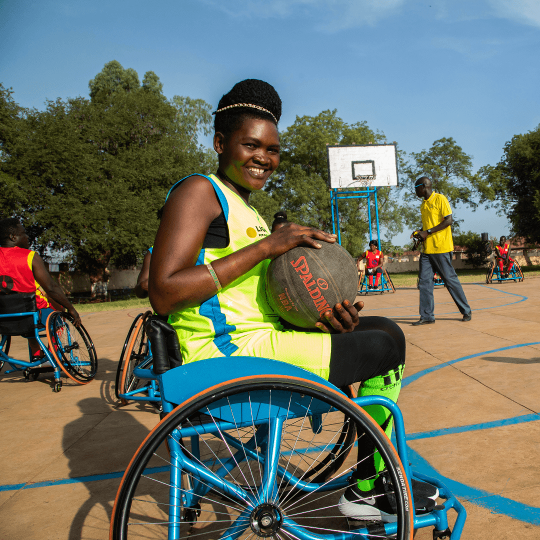 Eine junge Frau im Rollstuhl lächelt und hält einen Basketball. Sie trägt ein Basketballtrikot und befindet sich auf einem Basketballfeld.