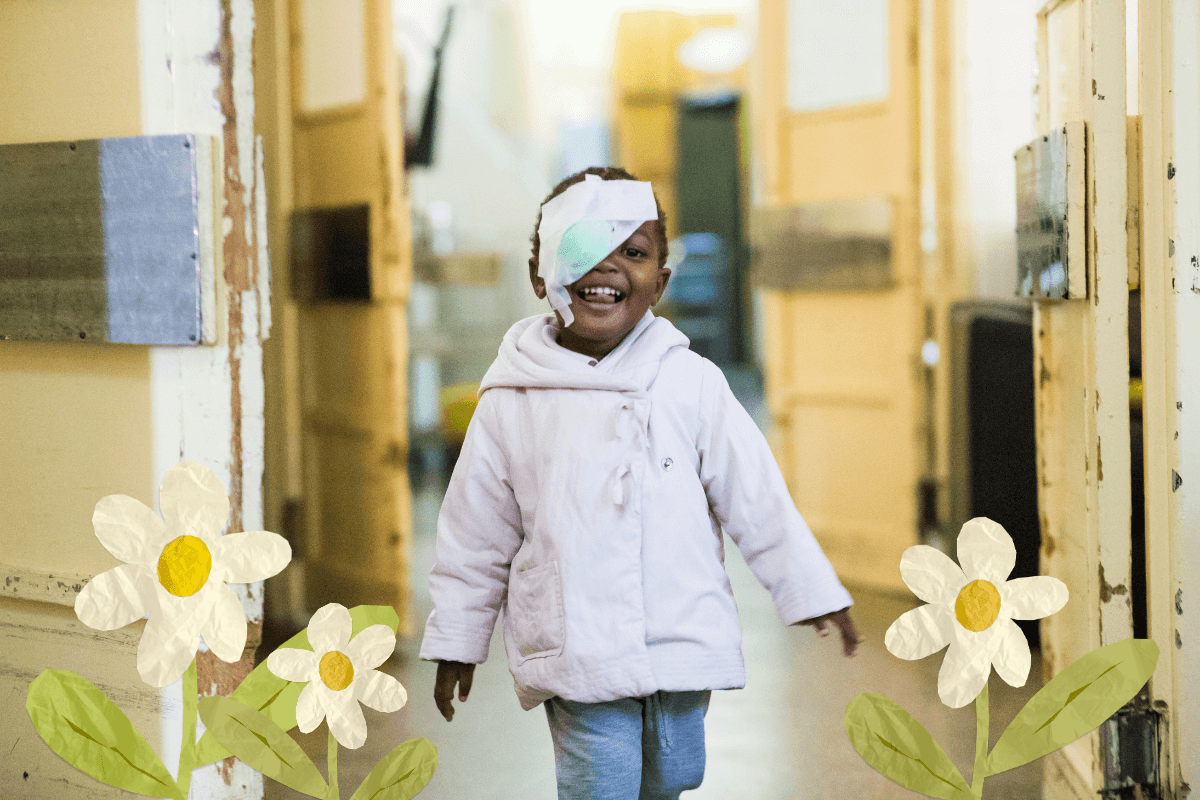 Ein kleines Mädchen mit Augenklappe lächelt nach ihrer erfolgreichen Kataraktoperation. Frühlingsblumen zieren das Foto.
