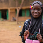 Das Bild zeigt ein Mädchen aus Burkina Faso. Sie trägt eine Schultasche und hat ein Tuch mit Perlenstickerein um den Kopf geschlungen.