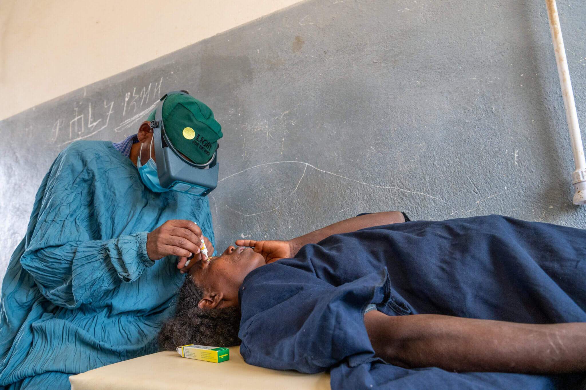 Auf dem Bild ist ein Chirurg in blauer Operationskleidung zu sehen. Auf dem Tisch liegt eine Frau, sie leidet an der Augenkrankheit Trachom. Der Arzt verabreicht ihr gerade Augentropfen.