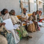 Auf dem Bild sind 6 Frauen zu sehen. Sie sitzen auf einer Bank und scheinen in ein Gespräch vertieft. In ihrer Hand halten sie ein Blatt Papier, ein Anmeldeformular. Sie sind für eine Augeuntersuchung gekommen.