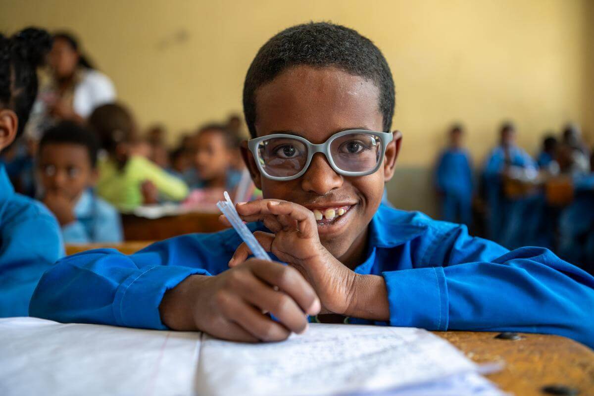 Der junge Kedir auf dem Bild grinst glücklich in die Kamera. Er sitzt in seinem Klassenzimmer und schreibt gerade in sein Schulheft. Er trägt seine neue Brille die er bekommen hat, um wieder klar sehen zu können. Ein wichtiger Schritt für gesunde Augen!