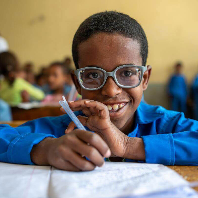 Der junge Kedir auf dem Bild grinst glücklich in die Kamera. Er sitzt in seinem Klassenzimmer und schreibt gerade in sein Schulheft. Er trägt seine neue Brille die er bekommen hat, um wieder klar sehen zu können. Ein wichtiger Schritt für gesunde Augen!