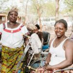 Zwei Frauen aus Burkina Faso lächeln in die Kamera. Eine von ihnen sitzt im Rollsthul und trägt ein weißes Shirt und einen bunten Rock. Die andere Frau steht daneben ebenfalls mit einem weißen Shirt und einem bunten Rock.