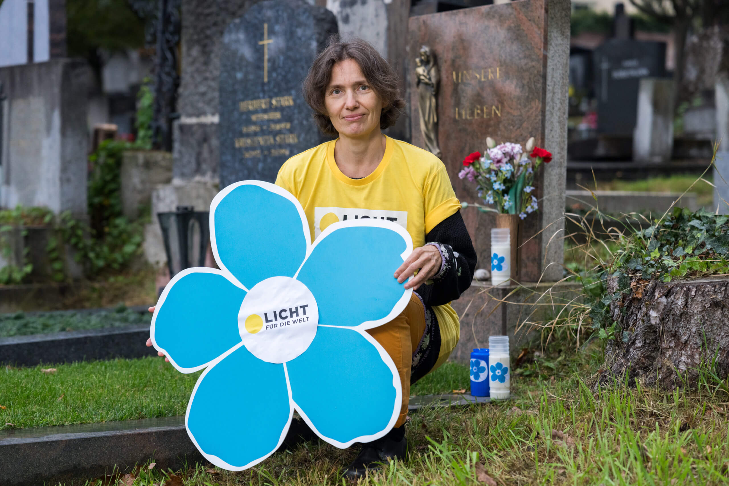 Eva Dürr im gelben Licht für die Welt T-Shirt mit einem großen blauen Vergissmeinnicht aus Karton in der Hand in der Hocke vor einem Grabstein