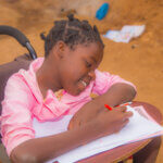 A girl in a wheelchair is writing on a piece of paper. The girl smiles.