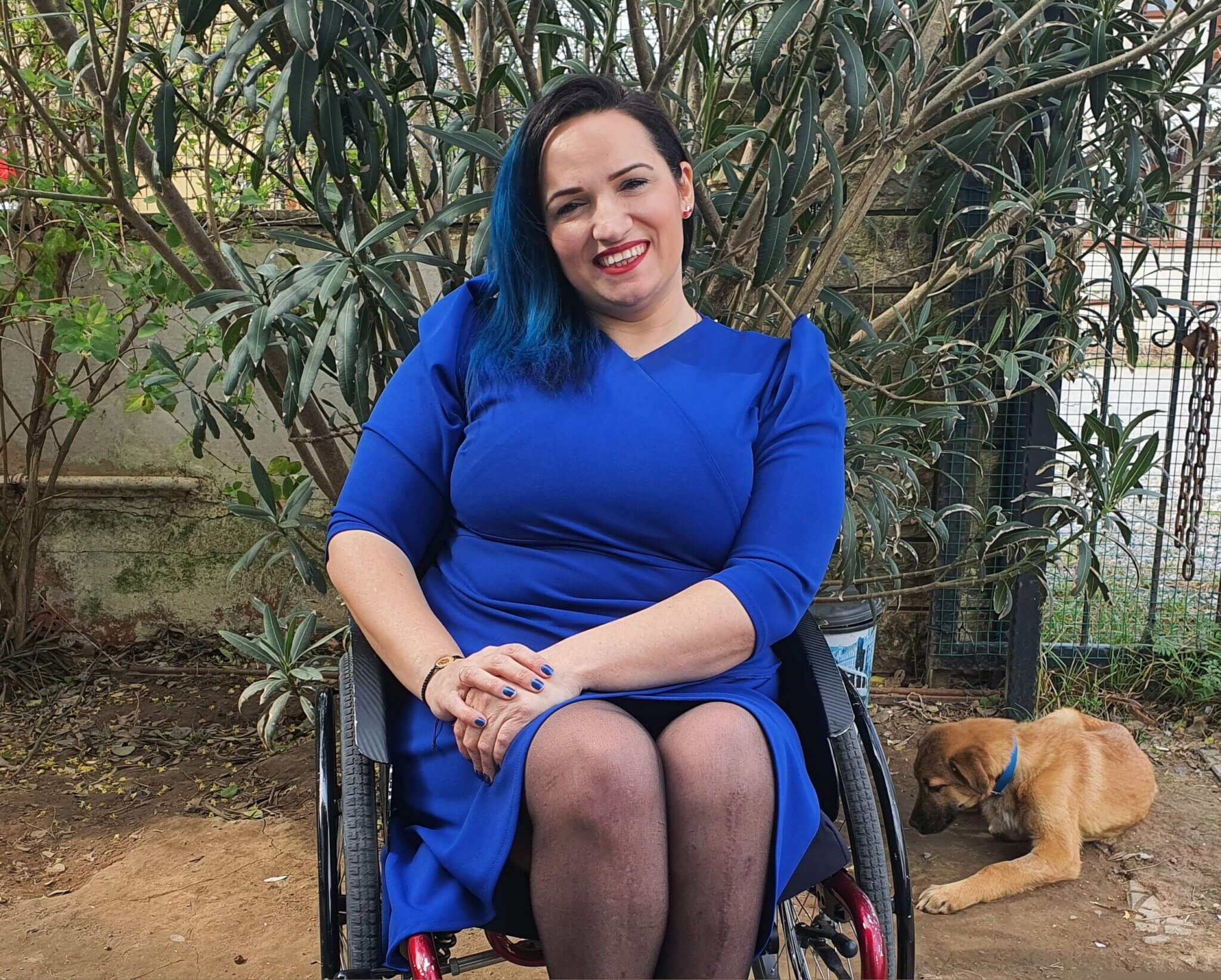 Die Regisseurin Gamze Elibol sitzt in einem blauen Kleid in ihrem Rollstohl. Die Hände hat sie auf dem linken Knie übereinandergelegt und lächelt in die Kamera.