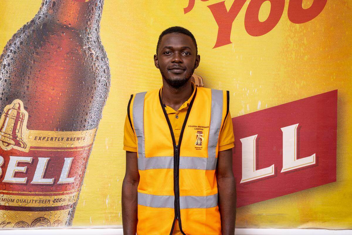 Higenyi Stuart Cyprian, ein Absolvent des BSc Industrial Chemistry, steht vor einer Werbewand der Uganda Breweries Limited. Er trägt eine Warnweste, seine Arbeitskleidung.