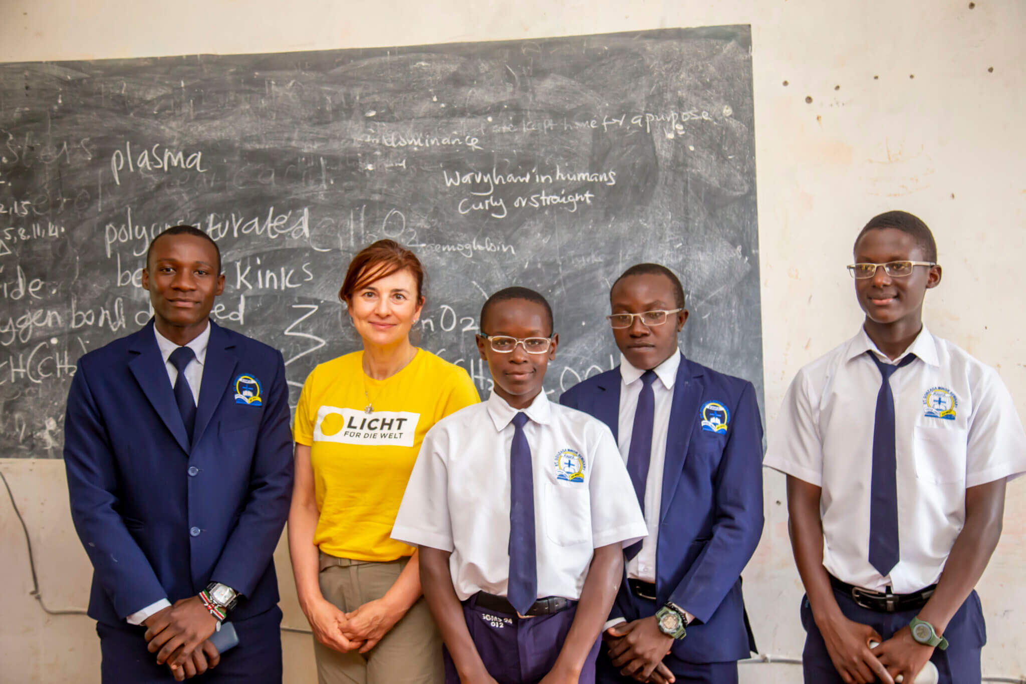 Auf dem Bild sind Augenärztin Irene Ruhswurm und Schüler der Jungenschule in Uganda zu sehen. Sie posieren vor einer Schultafel und lächeln in die Kamera.