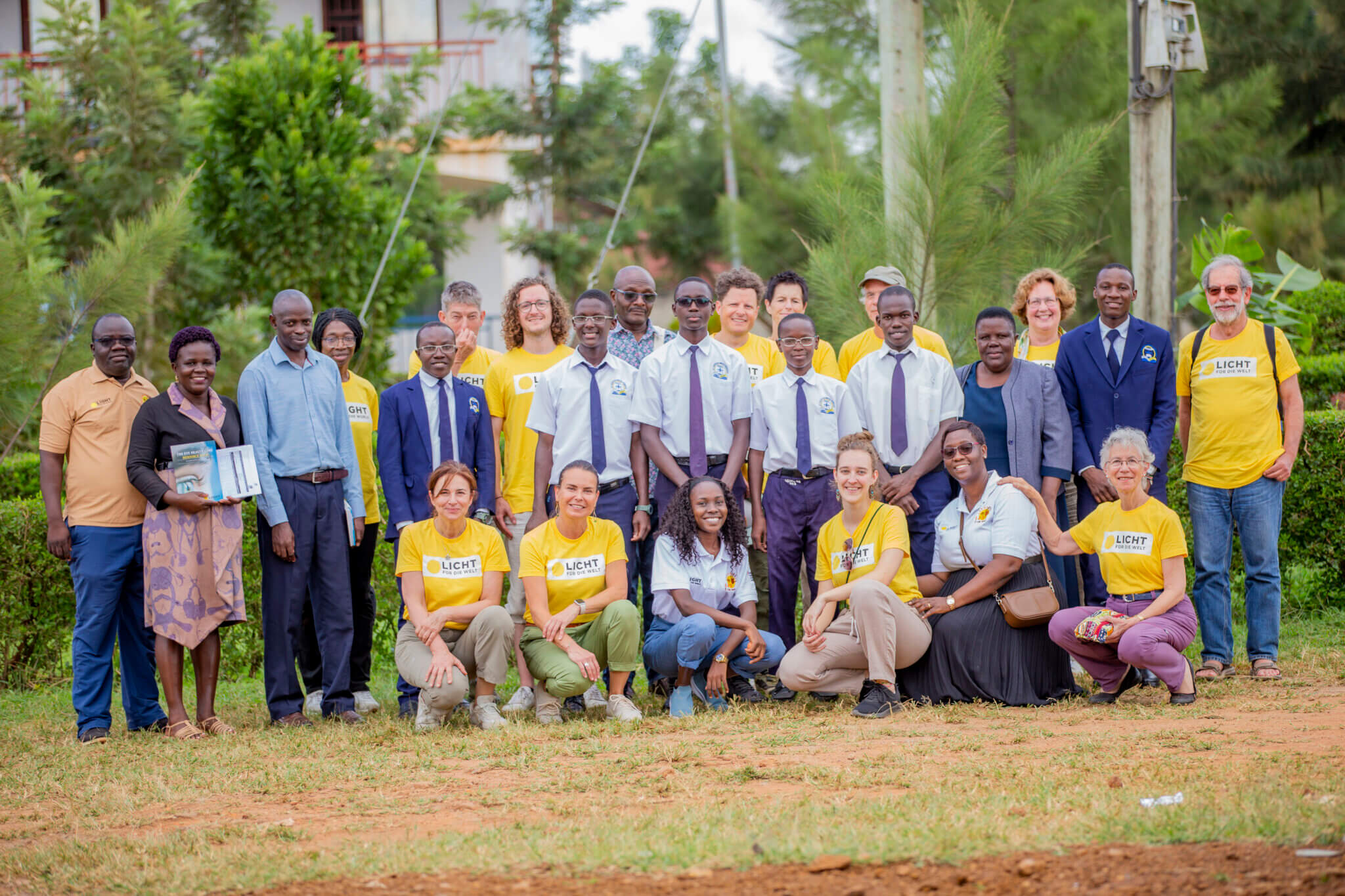 Auf dem Bild sind Personen verschiedenen Alters, Geschlechts und Hautfarbe zu sehen. Sie posieren für ein Bild, das im Rahmen der Reise von Augenärztinnen und Augenärzte mit Licht für die Welt nach Uganda aufngenommen wurde. Im Hintergrund ist dichtes Grün zu erkennen.