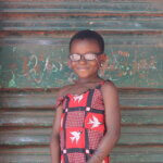Serena trägt ein rot-weiß gemustertes Kleid und eine Brille. Das Mädchen steht vor einer grünen Tür in ihrer Schule. Die Brille hat sie nach einer Schuluntersuchung von Licht für die Welt bekommen.