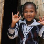 Die achtjährige Betselot aus Äthiopien steht vor der Lehmhütte in der sie mit ihrer Familie lebt. Sie hat die Hände gehoben und die Finger zu zeichen der Gebärdensprache geformt. Sie trägt eine gestreivte Weste und lächelt in die Kamera.