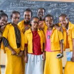 Nigest aus Äthiopien steht mir ihren Klassenkamerad*innen vor einer beschriebenen Tafel in ihrem Klassenzimmer und lachen in die Kamera. Alle tragen eine gelbe Bluse.