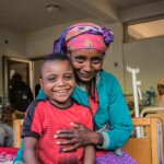 A woman with a colourful headscarf and a turquoise jacket is sitting on a hospital bed. She has her 8-year-old son in her arms, who has just survived a cataract operation. Both are smiling into the camera.