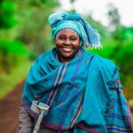 Lucy Nkatha, gekleidet in heller Tracht und mit einer Krücke in der Hand, lächelt.