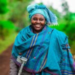 Das Bild zeigt Lucy Nkatha aus Kenia. Sie trägt einen blauen Schal und hat ein blaues Tuch um ihren Kopf geschlungen. Sie hält eine Krücke in der Hand und lächelt breit in die Kamera.