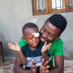 Das Bild zeigt den zweijährigen Aynir aus Mosambik. Er klatscht in die Hände. Hinter ihm kniet sein Vater und lächelt ihn an.