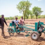 Das Bild ist auf einem leeren Feld aufgenommen. Eine Frau mit einer Behinderung sitzt auf einem Pluggerät, während zwei schwarze Männer neben ihr hergehen. Das Wetter ist sonnig, der Himmel blau.