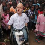 Tom Shakespeares ist umringt von Kindern aus Mosambik. Er sitzt im Rollstuhl und trägt ein weißes Hemd und blaue Jeans.