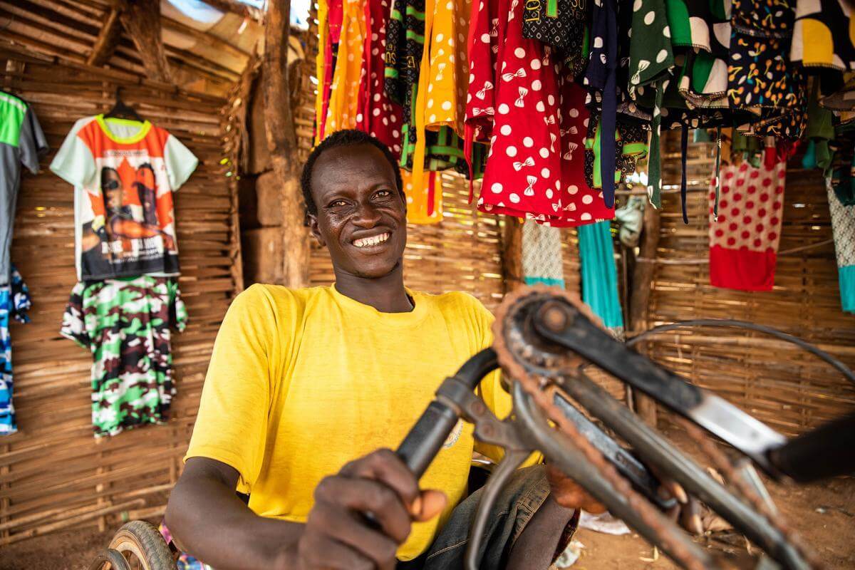 James Udodioy, ein Mann mit gelbem T-Shirt, sitzt auf seinem Tricycle in seinem Geschäft für Kindermode und lacht in die Kamera