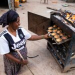 Eine Frau aus Uganda kniet vor einem Ofen und zieht mit der linken Hand ein Blech voller frisch gebackener Muffins aus dem Ofen.