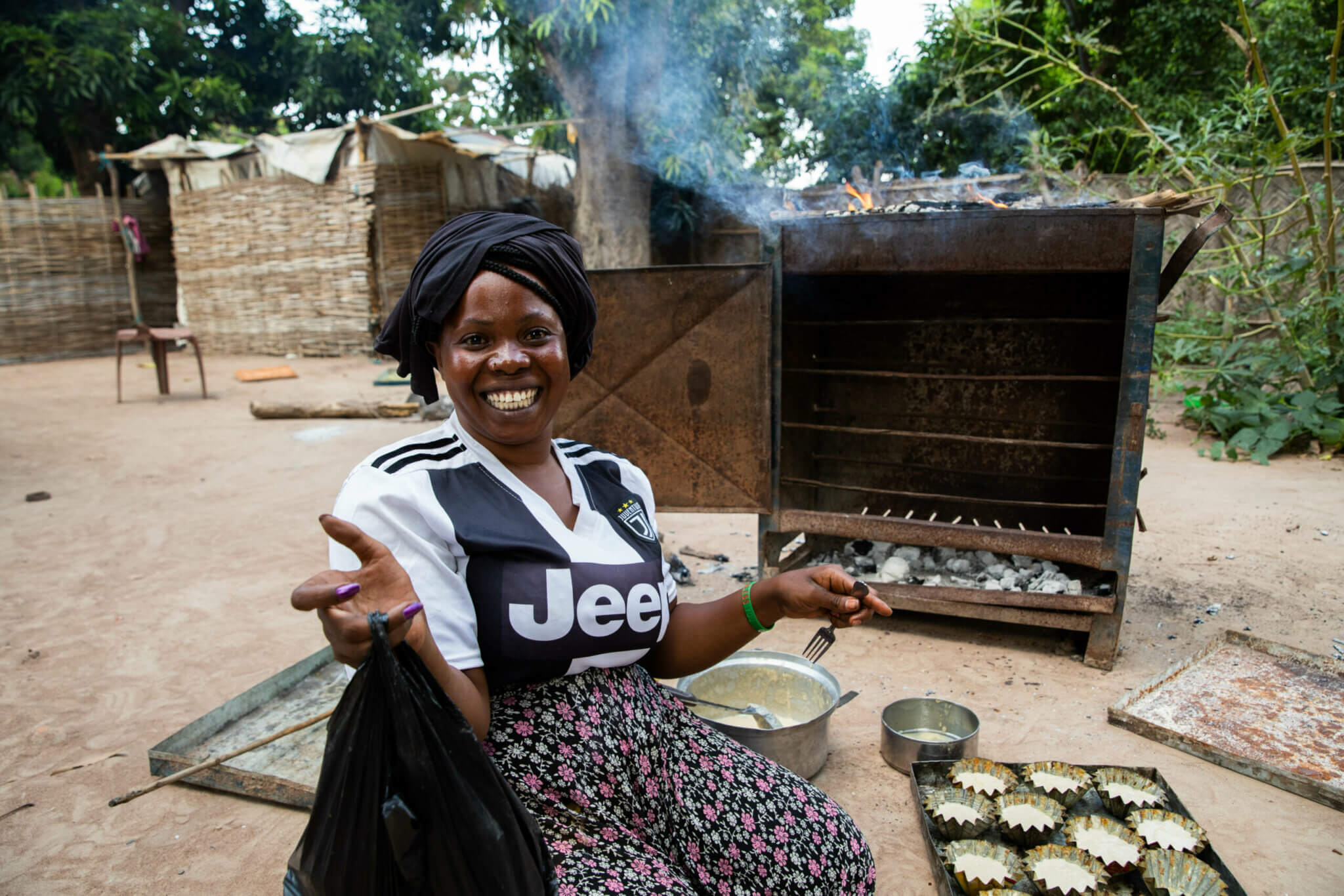 A woman with a black cloth on her head a white and black T-Shirt and a flowered skirt sits in her wheelchair and smiles into the camera. In the background you can see cakes in a form and a big oven.