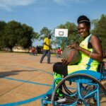 Lucy, eine Inklusionsberaterin von Licht für die Welt in Mosambik, sitzt im Rollstuhl und nimmt an einem Basketballtrunier teil. Sie trägt ein gelbes Trikot und hält den Ball lächelnd in ihren Händen.