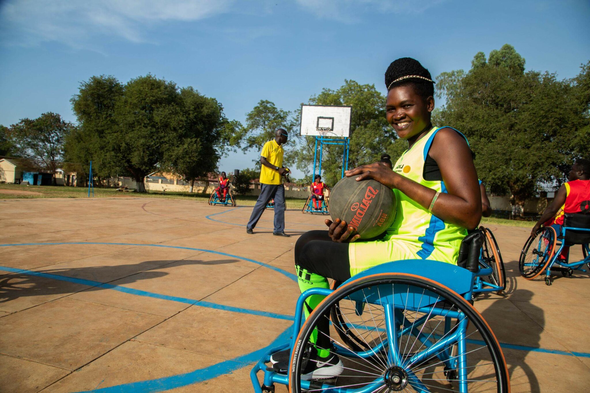 Lucy, eine Inklusionsberaterin von Licht für die Welt in Mosambik, sitzt im Rollstuhl und nimmt an einem Basketballtrunier teil. Sie trägt ein gelbes Trikot und hält den Ball lächelnd in ihren Händen.