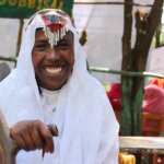 Das Foto zeigt eine Frau aus Afrika. Sie trägt ein traditionelles weißes Gewand mit bunten Perlen am Kopf und lächelt breit in die Kamera.