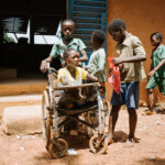 Kinder vor einer Schule und Junge schiebt den Rollstuhl eines Mädchens ohne Unterarme und Unterschenkel